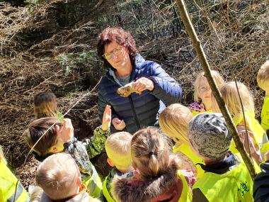 Pædagog holder dyrekranie i hånden og viser til børn inde i en skov